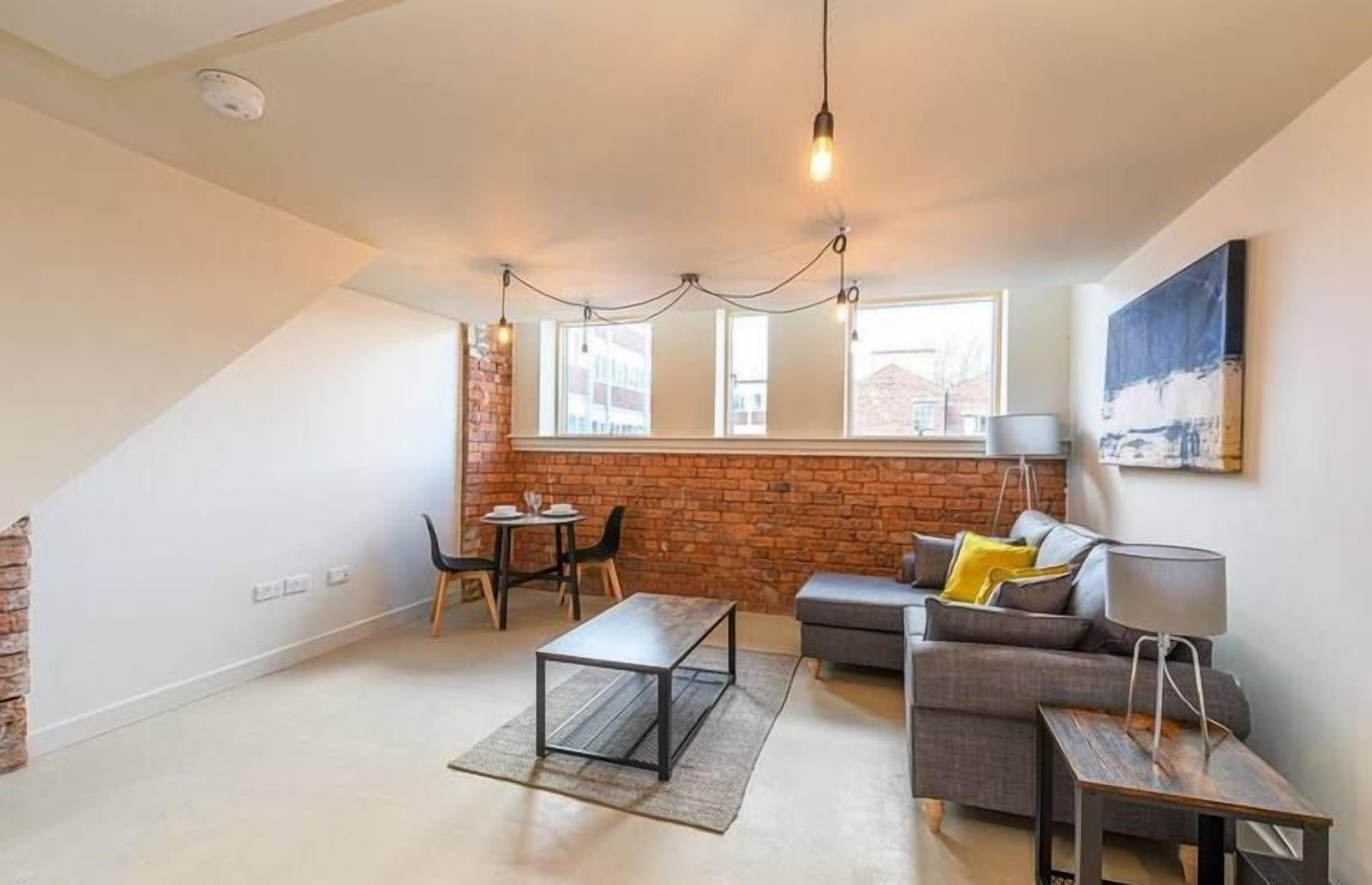 A modern living room with a sofa, coffee table, and dining area. There are large windows and a brick wall.