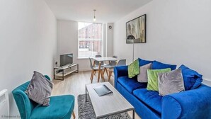 A modern living room with a blue sofa, a small table, a TV, and a dining area with chairs and a table. - Winkley House by Guestz (Preston)