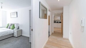 A modern bedroom with a bed, bedside table, and a mirror. A hallway leads to a kitchen area with cabinets and a microwave. - Winkley House by Guestz (Preston)