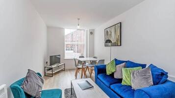A modern living room with a blue sofa, a small table, a TV, and a dining area with chairs and a table.