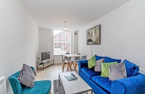 A modern living room with a blue sofa, a small table, a TV, and a dining area with chairs and a table. - Winkley House by Guestz (Preston)