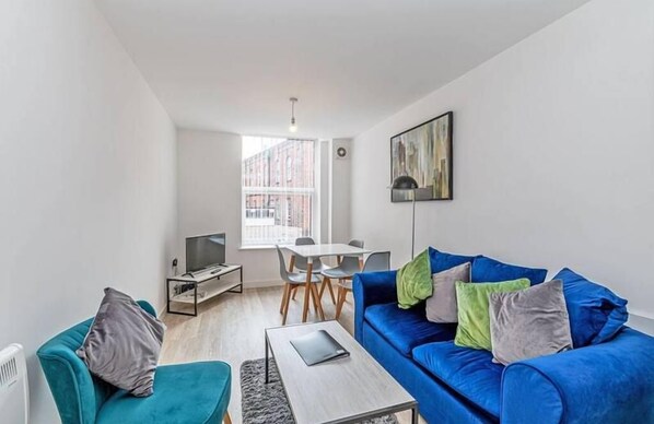 A modern living room with a blue sofa, a small table, a TV, and a dining area with chairs and a table.