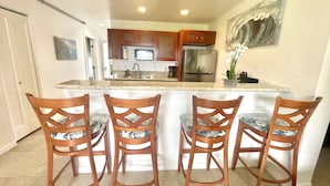 A kitchen with wooden cabinets, a stainless steel refrigerator, and a marble countertop. There are four wooden chairs with cushioned seats.