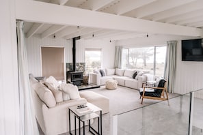 A spacious living room with a fireplace, large windows, and a flat-screen TV. - Ocean Paradise in Wine Country (Rarangi)
