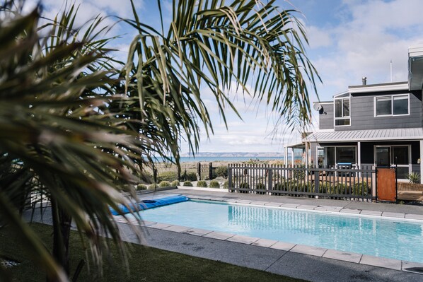 A swimming pool with a view of the ocean, surrounded by a fence and palm trees. - Ocean Paradise in Wine Country (Rarangi)