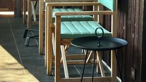 Een houten zitgedeelte buiten met een tafel en stoelen, een groen kussenbankje en een houten kastje met een glazen blad.