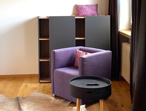 Un salon moderne avec un fauteuil violet, une table d’appoint noire et une bibliothèque intégrée sur laquelle repose un coussin rose.