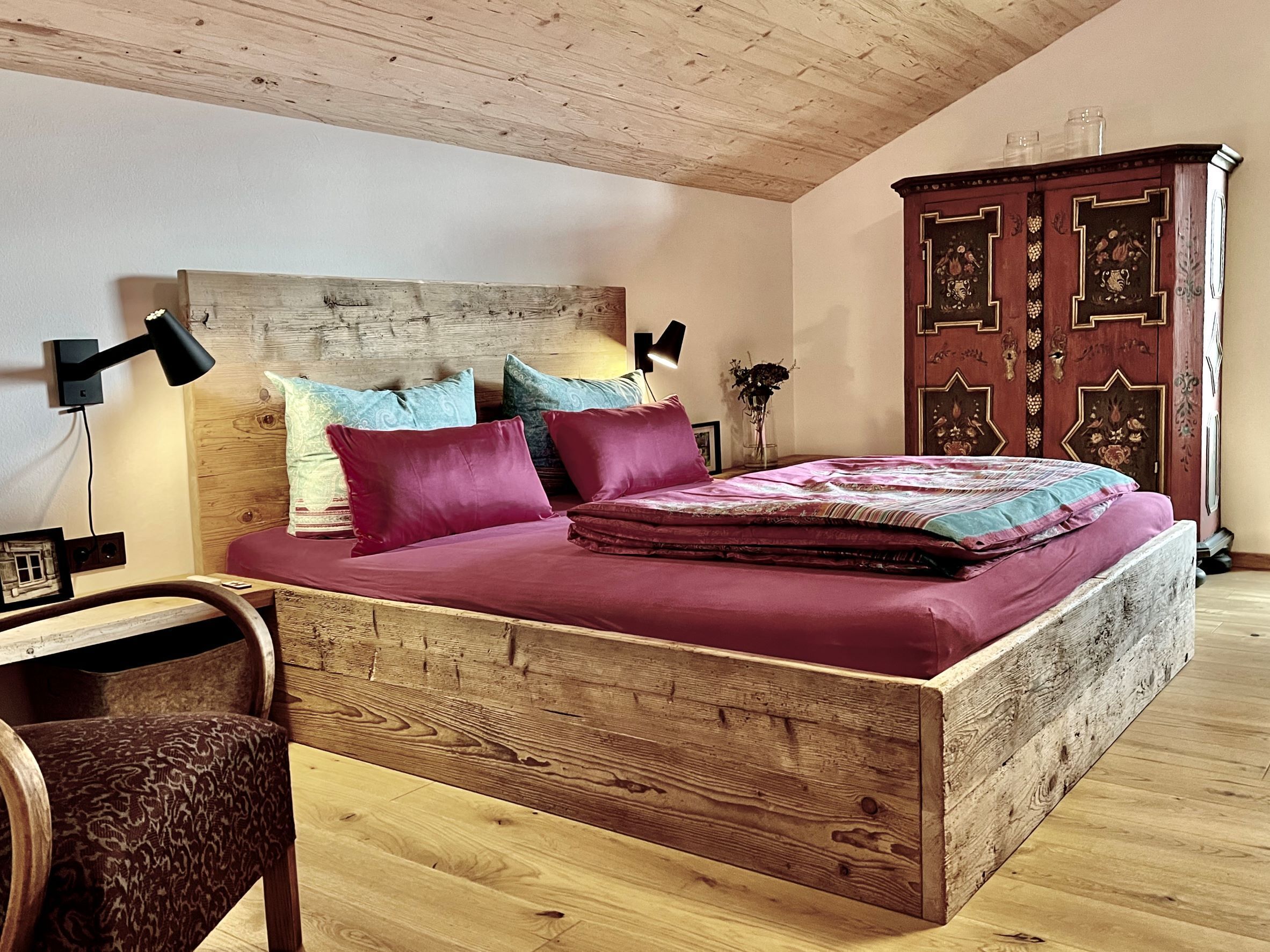 A bedroom with a wooden headboard, a bed with a red bedspread, two pillows, a nightstand with a lamp, and a wooden cabinet.