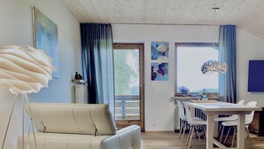 Een moderne woonkamer met een bank, een eettafel en stoelen. Er is een groot raam met gordijnen, kunstwerken aan de muur en een vloerlamp.