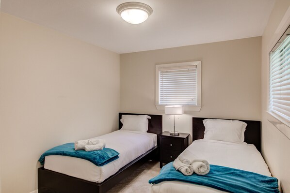 Une chambre d’hôtel avec deux lits simples, une table de chevet avec une lampe, une fenêtre avec des stores et un luminaire au plafond.