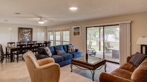 A spacious living room with a blue sofa, a tan armchair, and a glass coffee table. There is a dining area with a table and chairs, and a large window offering a view of the outdoors. - 🌵 Austin Escape with Private Pool, Ping Pong & Game Space | Roadrunner Retreat (Austin)