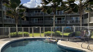 A swimming pool with a stone tile border, surrounded by palm trees and a multi-story building with balconies. - Cozy Studio By The Beach! 1BR (Kapaʻa)