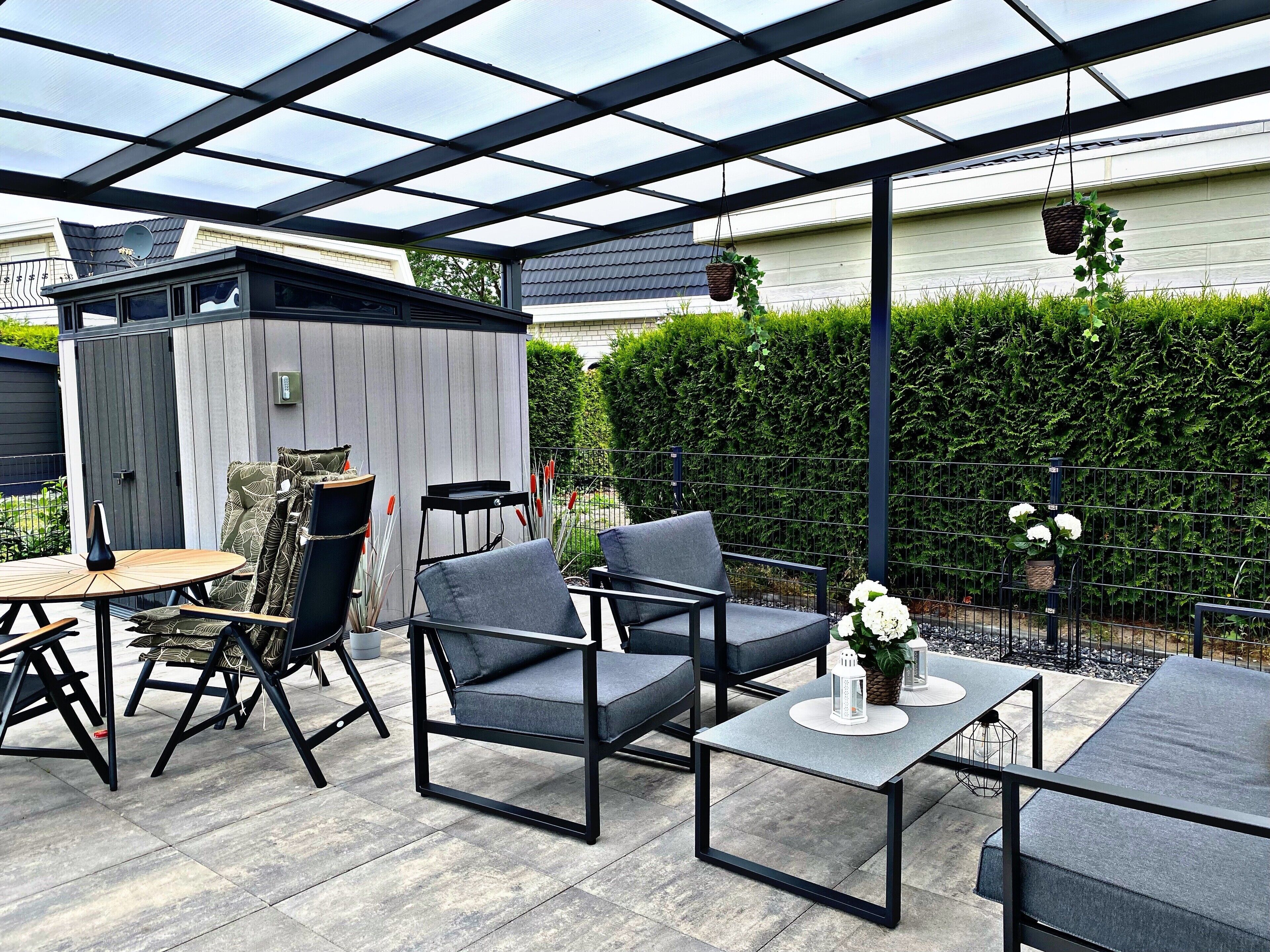 A covered patio area with a table and chairs, a small shed, and a hedge in the background.