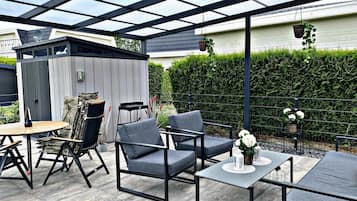 A covered patio area with a table and chairs, a small shed, and a hedge in the background.