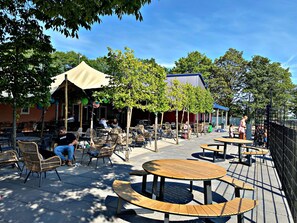 Un espace extérieur aménagé en terrasse, avec des tables rondes en bois et des chaises en osier, entouré d’arbres et bordé d’un bâtiment dont le toit est bleu.