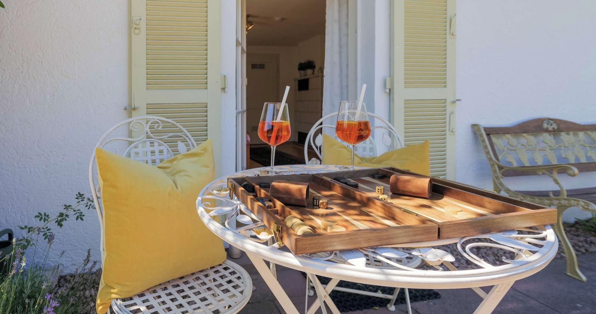 A round metal table with a wooden tray, two yellow cushions, and two wine glasses with red liquid.