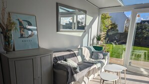 A modern living room with a grey sofa, a small white table, and a cabinet. There is a framed artwork on the wall and a window overlooking a garden. - Spacious house 200m from Lilia's beaches & restaurants (Plouguerneau)