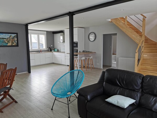 Ein modernes Wohnzimmer mit einem schwarzen Ledersofa und einem blauen Sessel, einer Holztreppe und einer Küche im Hintergrund.