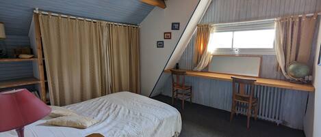 Une chambre avec un plafond en pente, un lit recouvert d’une couette blanche, un bureau en bois, deux chaises, un radiateur et une fenêtre avec des rideaux.
