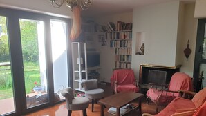 A living room with a fireplace, a bookshelf, and a large window with a view of greenery. - Large family home, between beaches and marshes. (Mesquer)