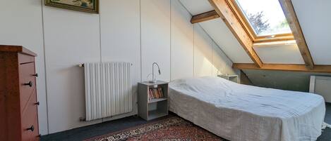 Un dormitorio con techo inclinado, tragaluz, radiador, mesita de noche con lámpara, cama con cubierta blanca, alfombra estampada y cómoda.