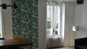 Una habitación con una mesa de comedor de madera, sillas, un sofá y una lámpara de pie. Hay una ventana con cortinas y un radiador.