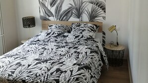 Un dormitorio con cama, un mural de hojas, una lámpara y una mesita.