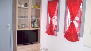 Une pièce comprenant un buffet en bois contenant de l’ustensile de cuisine, un téléviseur à écran plat et une petite table. Il y a deux fenêtres avec des rideaux rouges.