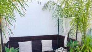 Een zithoek met een donkere wickerbank, witte kussens en een groene plaid. Aan weerszijden van de bank staan potplanten.
