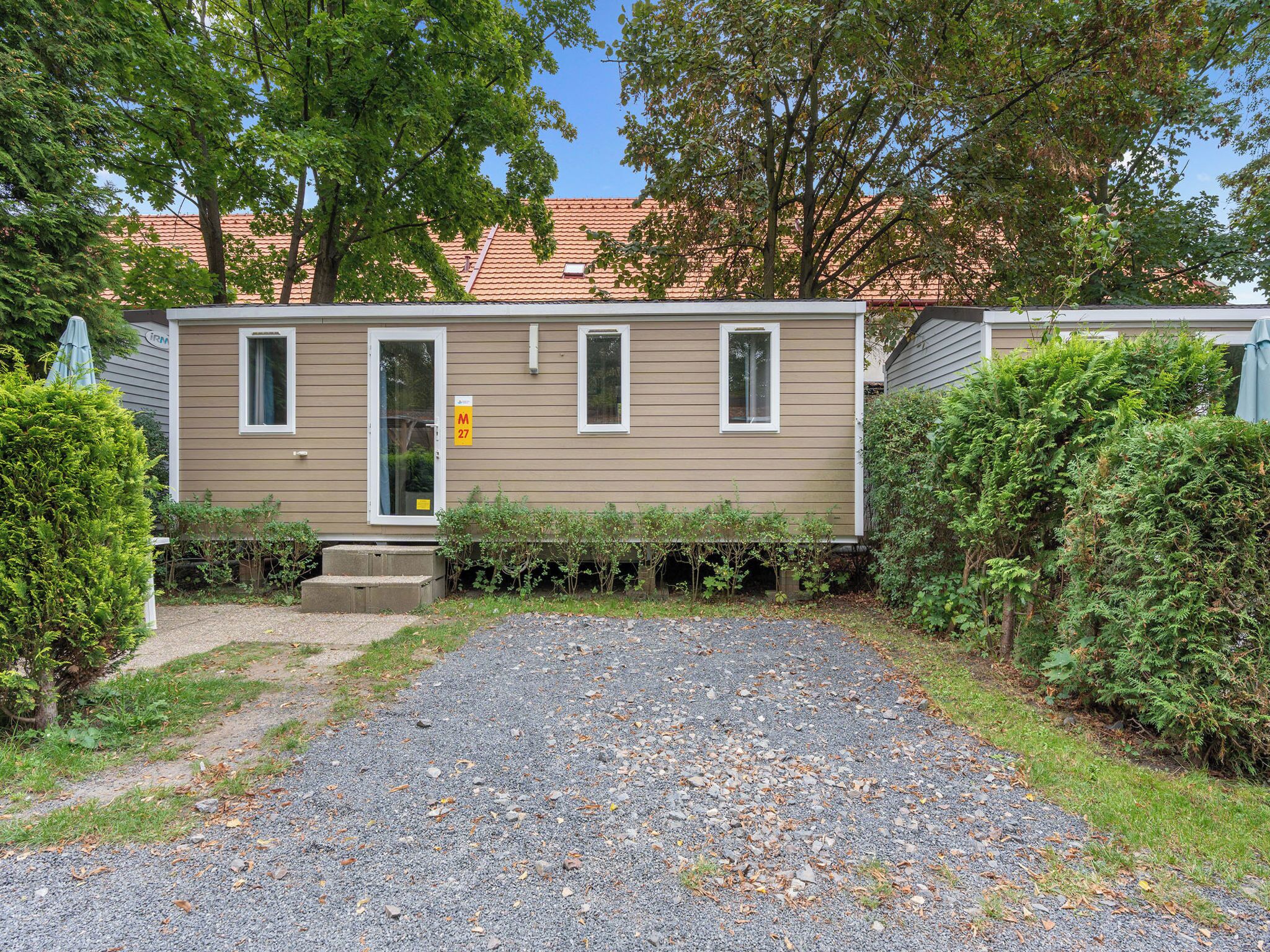 A single-story mobile home with a gravel driveway, surrounded by greenery and trees.