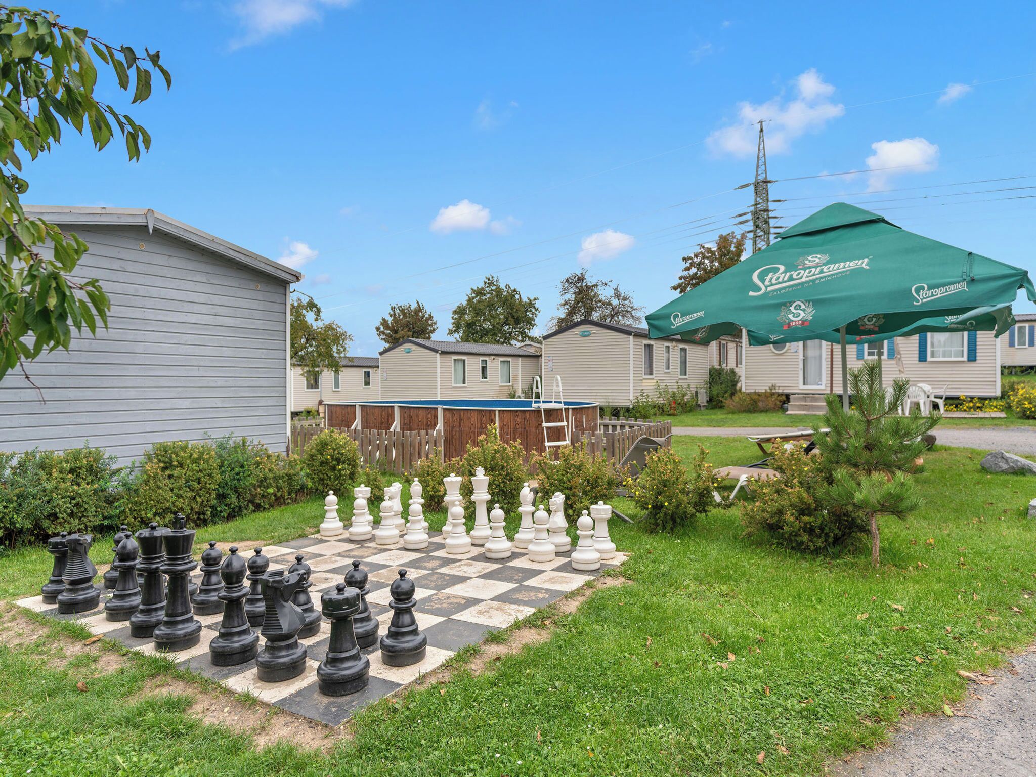 A chessboard set up in a grassy area with a large green umbrella and a wooden deck nearby.