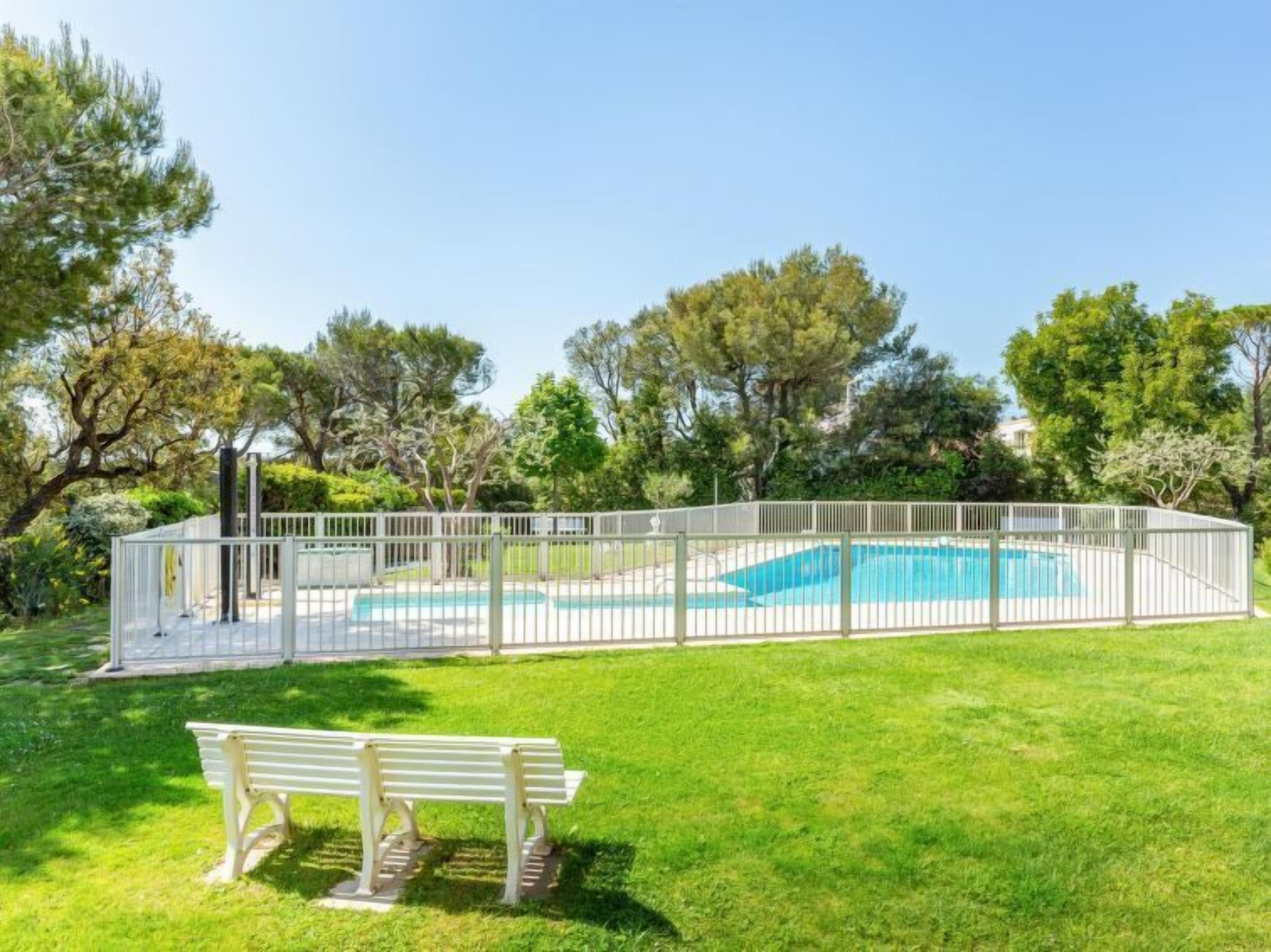 A rectangular swimming pool surrounded by a white fence, with a bench in the foreground and trees in the background.