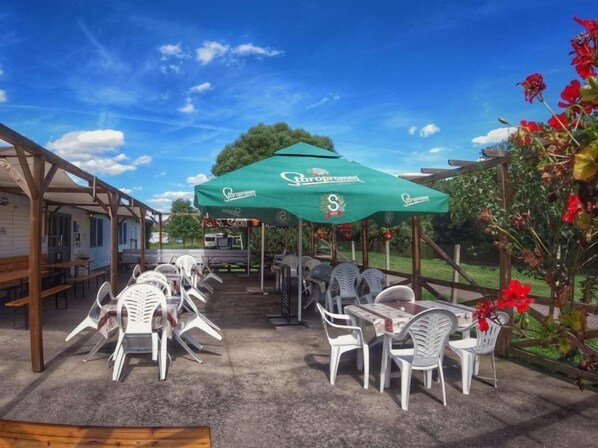 An outdoor patio with white plastic chairs and tables, green umbrellas, and a wooden pergola structure.