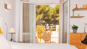 Ein Schlafzimmer mit Bett, Nachttisch, Lampe, Kommode und Blick auf einen Außenbalkon mit Tisch, Stühlen und Pflanzen.