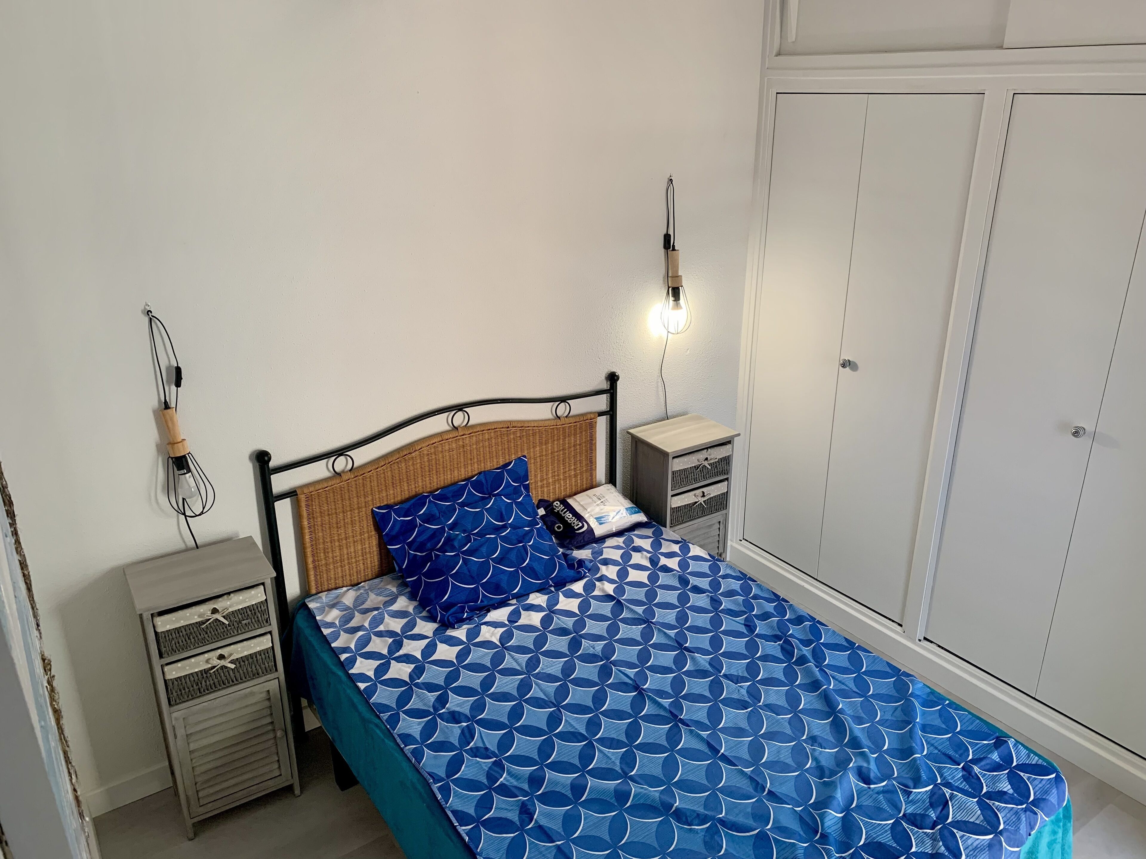 A neatly made bed with a blue patterned bedspread, a wicker headboard, and a single blue pillow. There are two bedside tables with lamps, a wardrobe, and a small cabinet with drawers.
