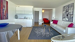 A modern living room with a white sofa, a dining table with a blue bowl, a kitchen area with white cabinets, and a red bed in the background. - Larmor-Plage 1BR with Terrace and Garage (Larmor-Plage)