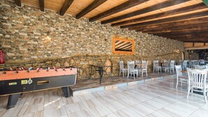 Un mur en pierre, des poutres en bois au plafond, une table de baby-foot et des chaises blanches dans une pièce.