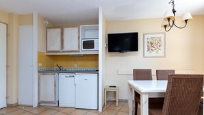 A compact kitchen with a microwave, a small refrigerator, a dining table with chairs, a television mounted on the wall, and a framed picture.