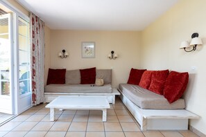 Un salon avec un canapé, une table basse blanche et un banc. Il y a deux lampes fixées au mur et un tableau encadré accroché au mur.