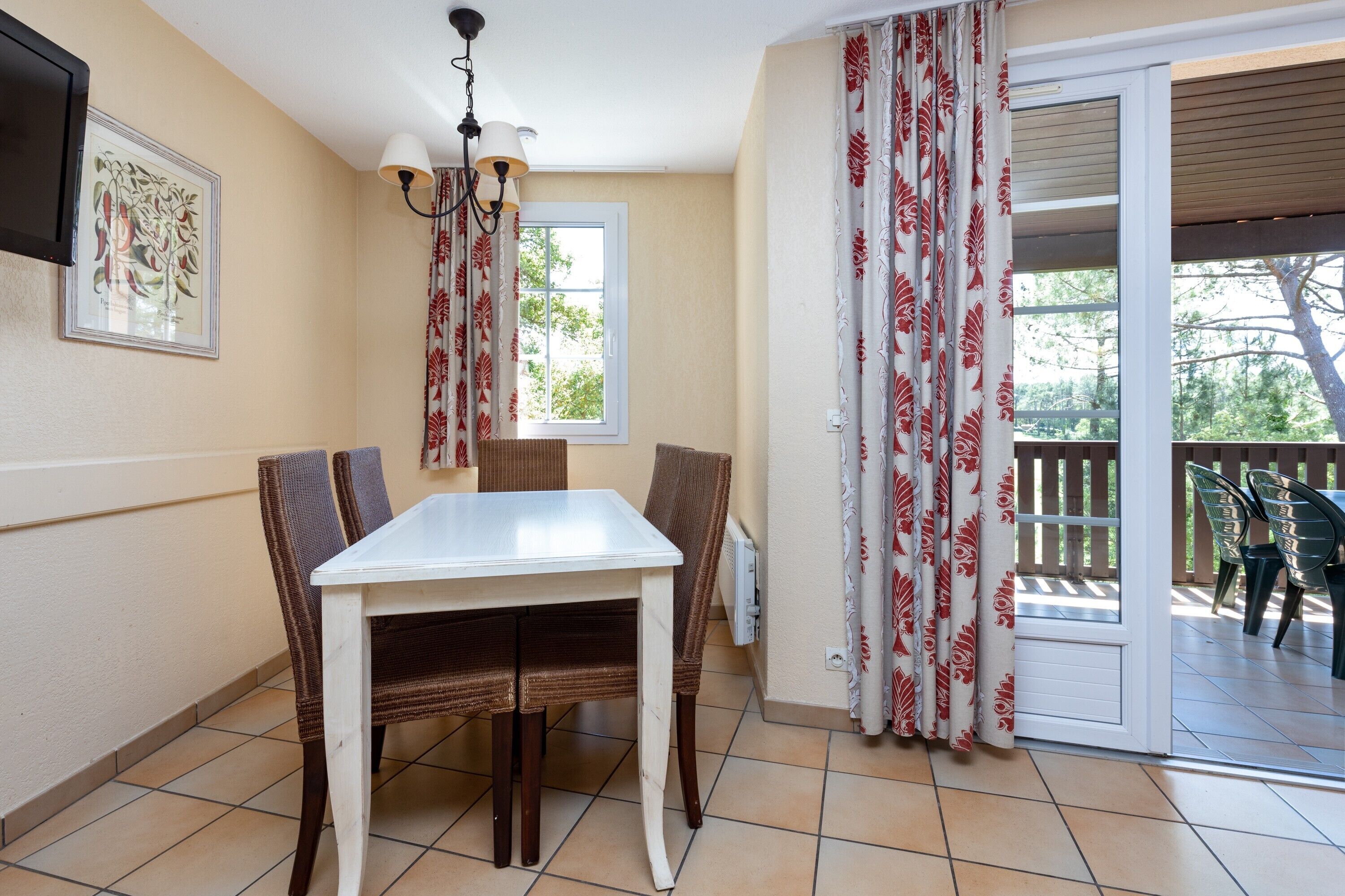 A dining area with a white table and wicker chairs, a chandelier, a window with curtains, and a view of an outdoor patio.