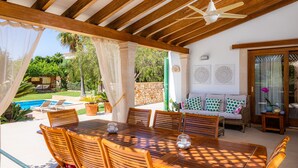Une terrasse extérieure avec une table et des chaises en bois, un ventilateur de plafond et une vue sur une piscine et la verdure.
