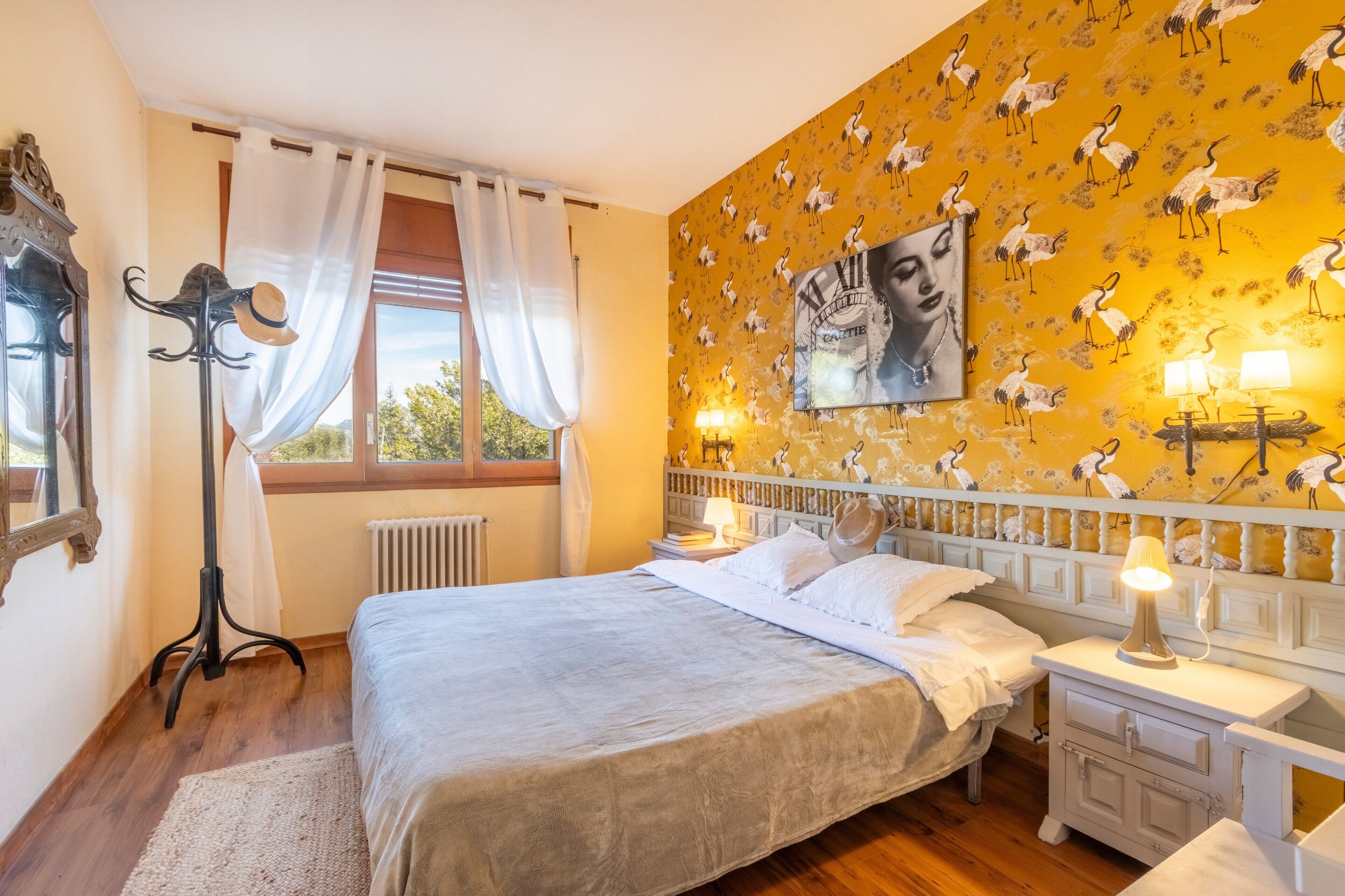 Un dormitorio con cama, mesitas de noche, lámpara de pie, radiador y ventana con cortinas.