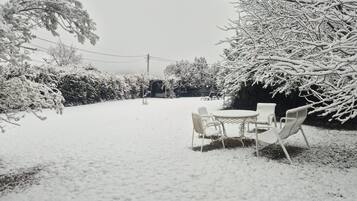 Un espacio exterior cubierto de nieve con una mesa y sillas, rodeado de árboles y arbustos cargados de nieve.
