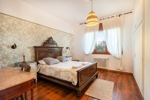 Un dormitorio con cama de madera, una mesita de noche, un escritorio, una ventana con cortinas y un radiador.