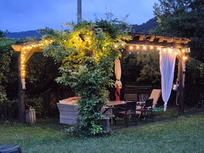 Un patio al aire libre con una mesa y sillas de ladrillo, luces colgantes y una pérgola cubierta de plantas trepadoras.