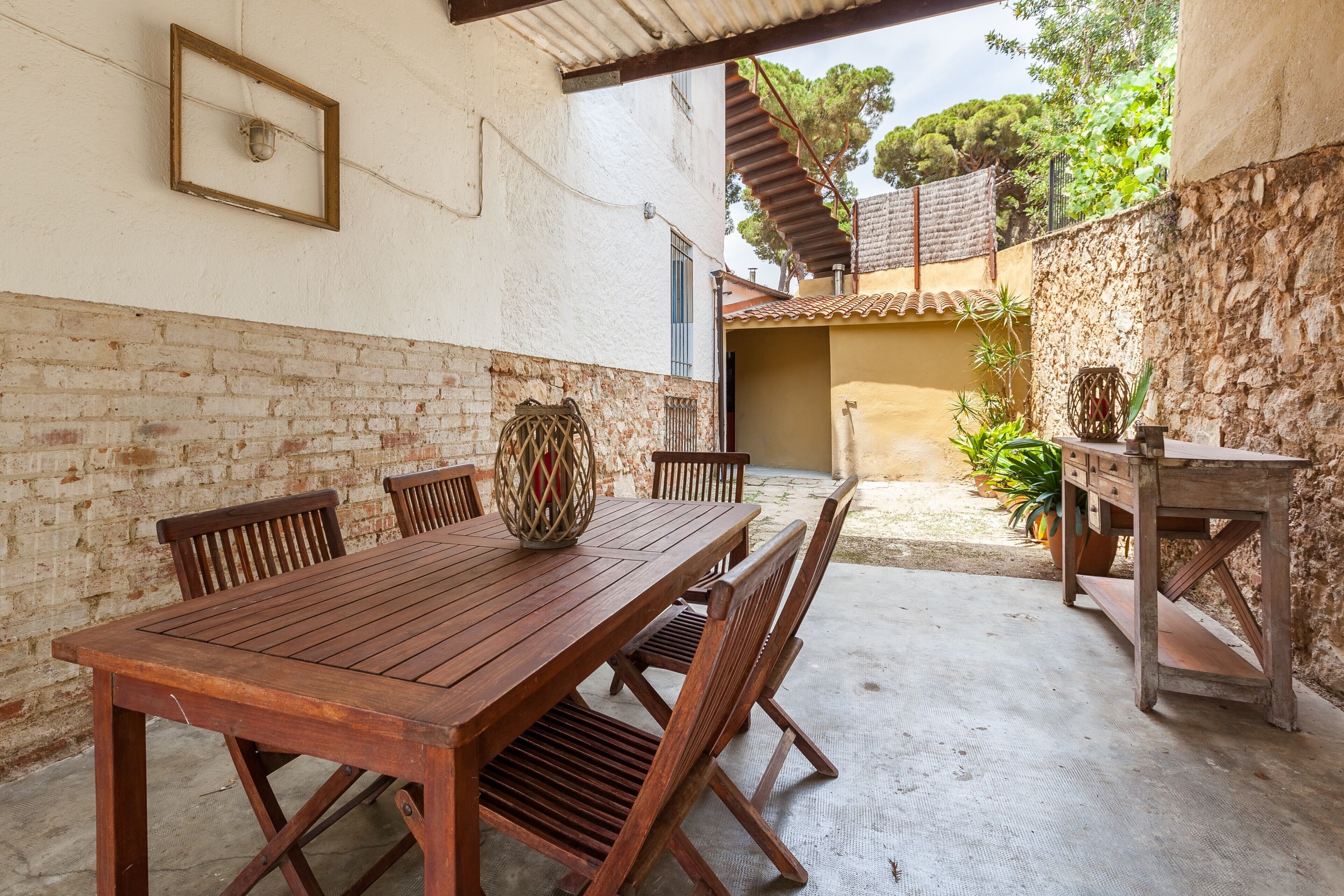 Una mesa y sillas de madera en un espacio exterior cubierto, con un muro de ladrillo y uno de piedra.