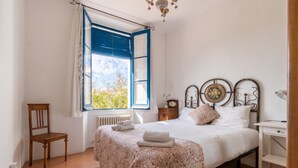 Een slaapkamer met een bed, een stoel, een nachtkastje, een raam met blauwe luiken en een radiator.