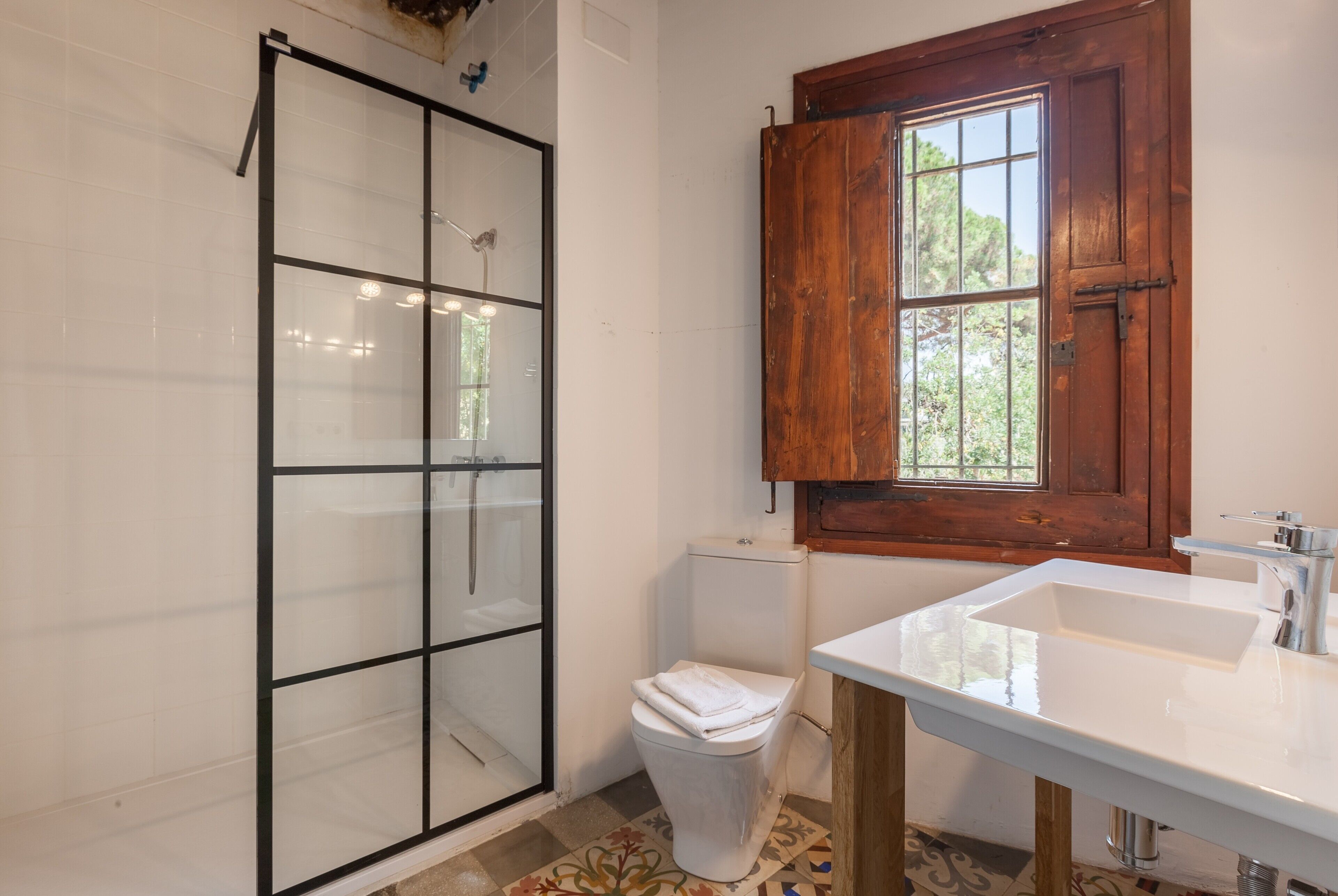 A bathroom with a glass shower enclosure, a white toilet, a wooden window, and a white sink.