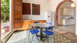 Een eetruimte met een glazen tafel en blauwe stoelen, een houten deur, en een keuken met witte kasten.