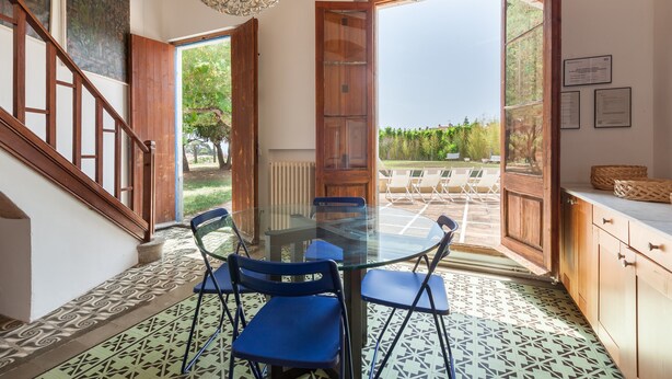 Een eetgedeelte met een glazen tafel en blauwe stoelen, een houten trap en uitzicht door openslaande deuren.